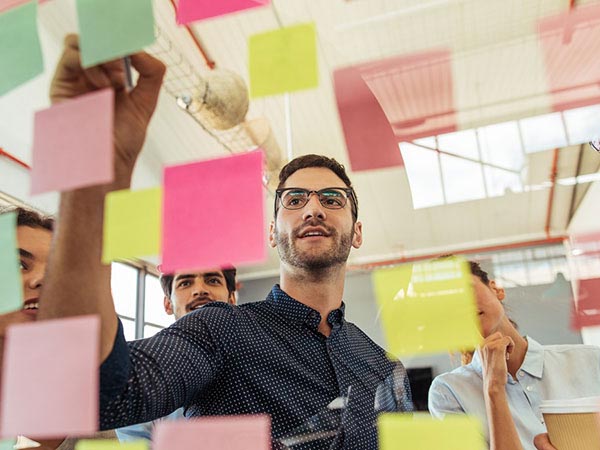 a man writing on colored post it notes on a transparent board with 3 people standing behind him