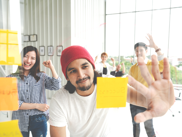 man looking at sticky note posted on glass wall while teammates cheer him on behind him