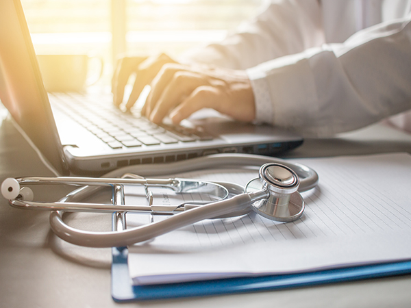 Healthcare provider using a laptop next to a stethoscope on top of a clipboard