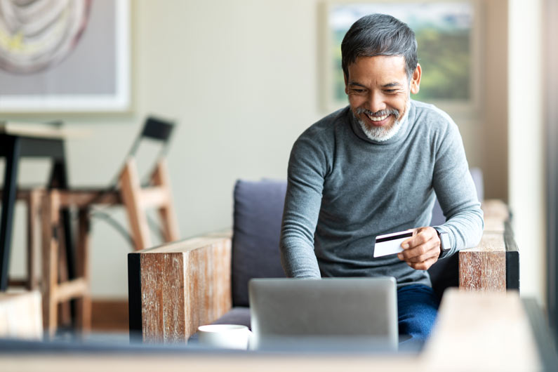 photograph of a man sitting in his home at a laptop while holding a credit card 